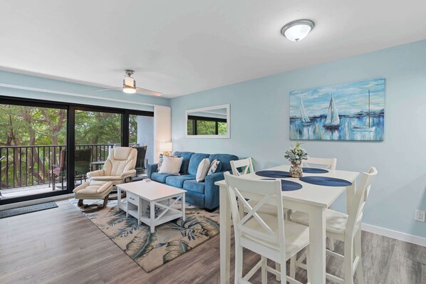A spacious living room with a blue sofa, a white dining table, and a ceiling fan.