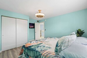 Quarto com cama, mesa de cabeceira, ventoinha no teto e porta que dá para outro cômodo.