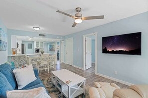 Sala de estar com um sofá azul, uma mesa de centro branca e um sofá bege. Há um ventilador de teto, uma pintura de uma noite estrelada e uma cozinha ao fundo.