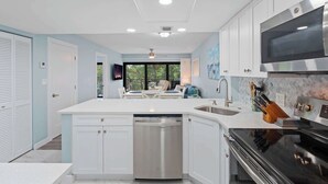 A modern kitchen with white cabinetry, a stainless steel dishwasher, a black stove, and a large window providing a view of the outdoors. - J-357 - Luxury With Private Lanai View & Sleeps 6 (Naples)