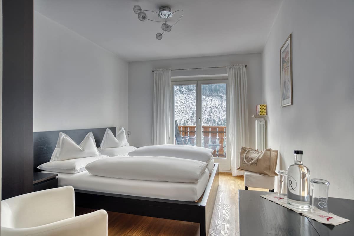 Hotellihuone, jossa on sänky, tuoli, pöytä, ikkuna verhoilla, patteri ja seinällä oleva kehystetty kuva.