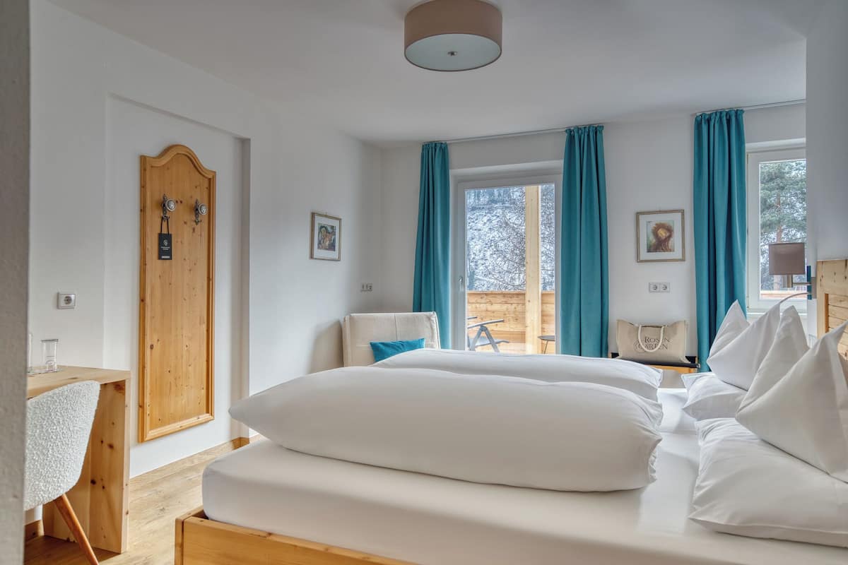 Hotellihuone, jossa on puinen ovi, valkoisilla lakanoilla varustettu sänky, työpöytä tuolilla ja suuri ikkuna, josta avautuu näkymä ulos, ja ikkunassa on siniset verhot.