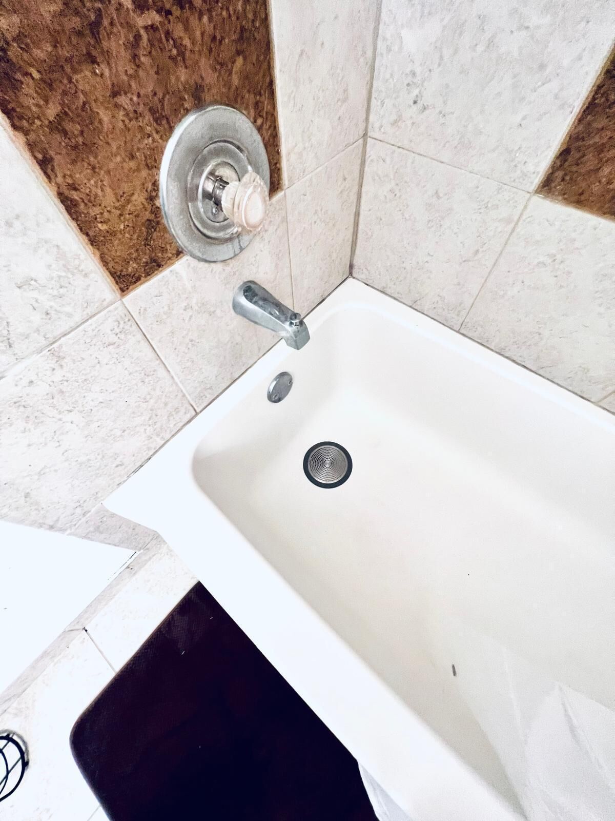 Une salle de bain avec une baignoire blanche, un robinet argenté et un drain. Les murs sont recouverts de carreaux de couleurs beige et marron.