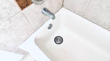 Une salle de bain avec une baignoire blanche, un robinet argenté et un drain. Les murs sont recouverts de carreaux de couleurs beige et marron.