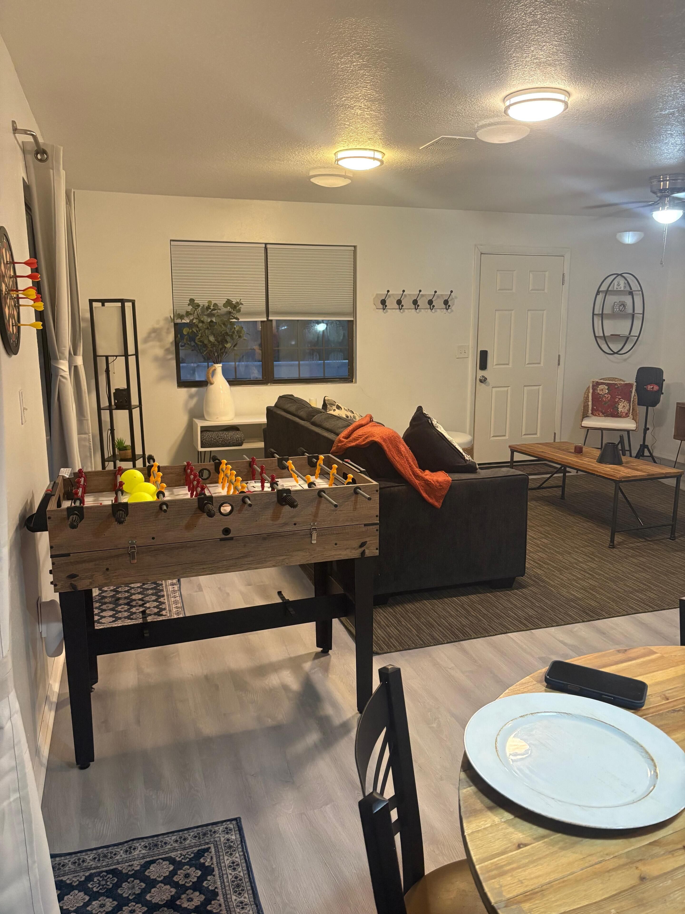 Uma sala de estar com mesa de futebol, sofá, mesa de jantar com cadeiras, mesa de centro, televisão e janela com persianas.