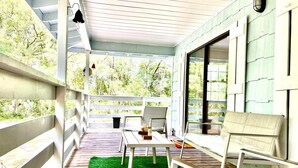 A covered patio with a table and chairs, a green artificial turf, and a view of trees through large windows. - Blue Sky StiltsHouse in a Secluded Forest. (Ocala)