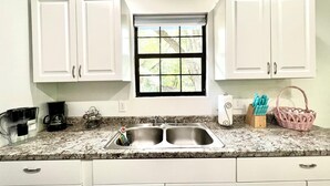 A kitchen with white cabinets, a granite countertop, a double sink, a coffee maker, and a window with a view of greenery. - Blue Sky StiltsHouse in a Secluded Forest. (Ocala)