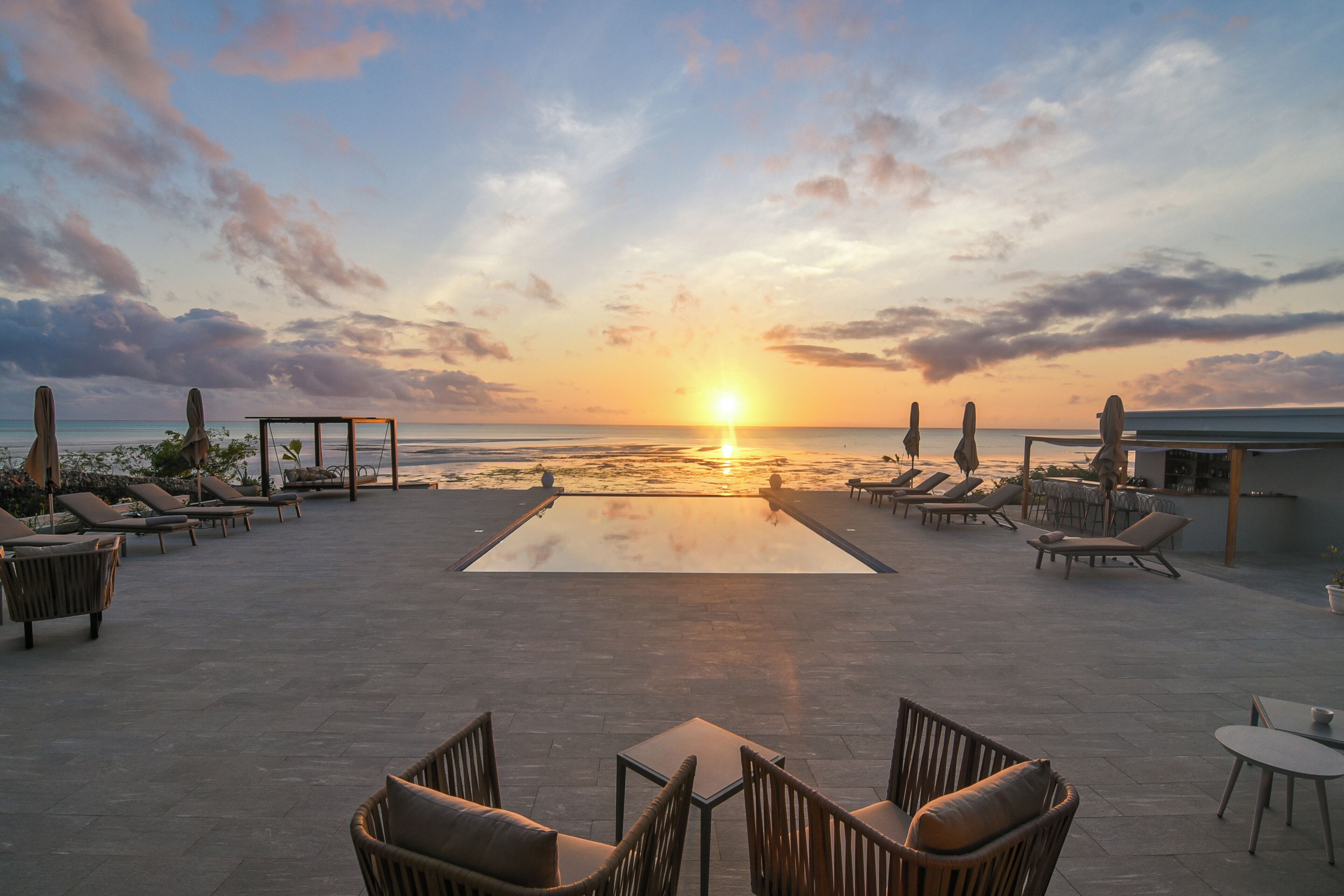 A rooftop terrace with lounge chairs and a pool, overlooking the ocean at sunset.
