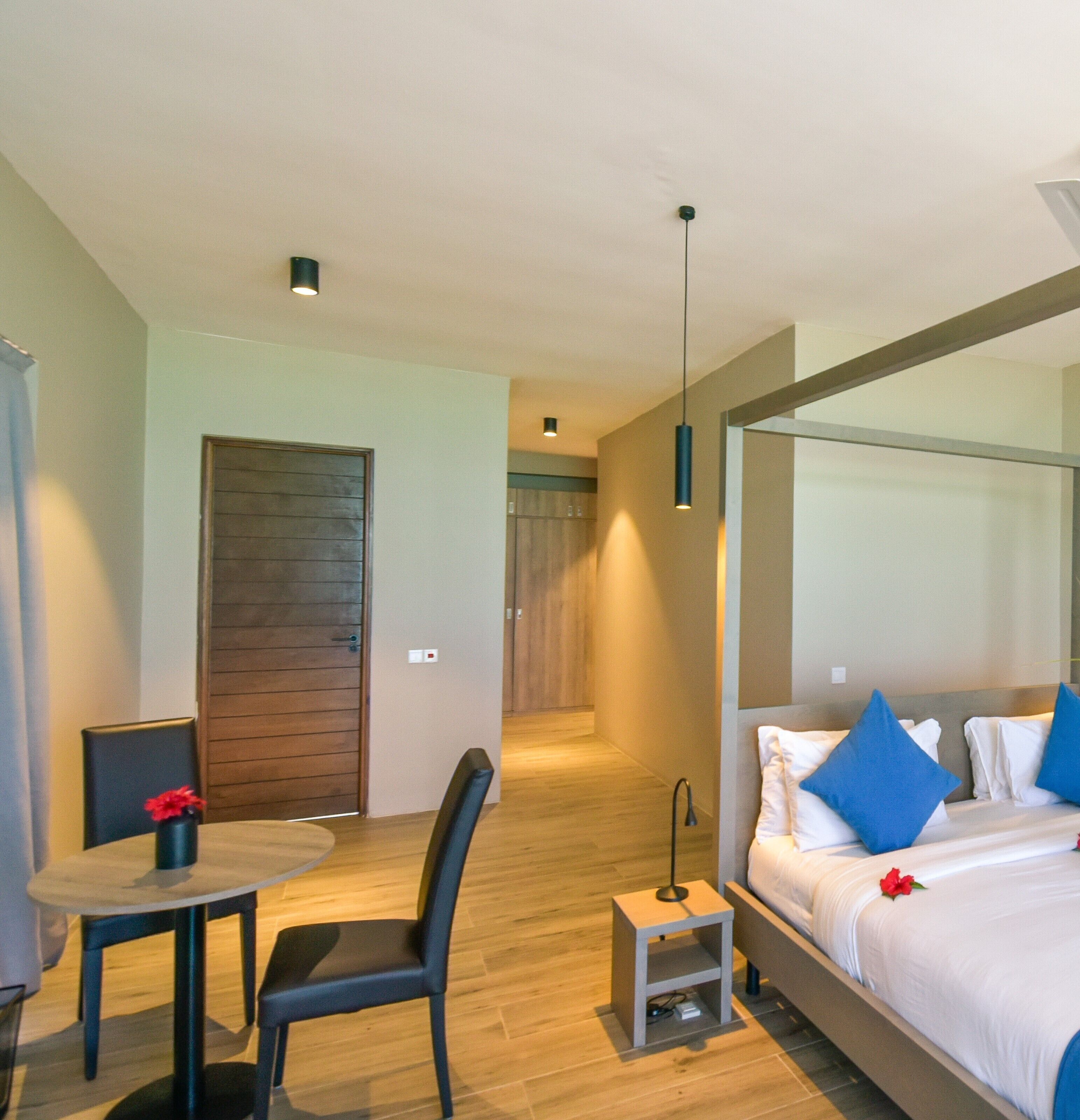 Une chambre d’hôtel moderne avec un plancher en bois, un lit avec une literie blanche et des oreillers bleus, une petite table avec une chaise, et une porte donnant sur une autre pièce.