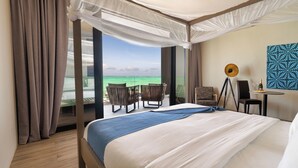 A hotel room with a large bed, a balcony with a view of the ocean, a desk with a chair, and a decorative wall piece. - Villa Bluu (Jambiani)