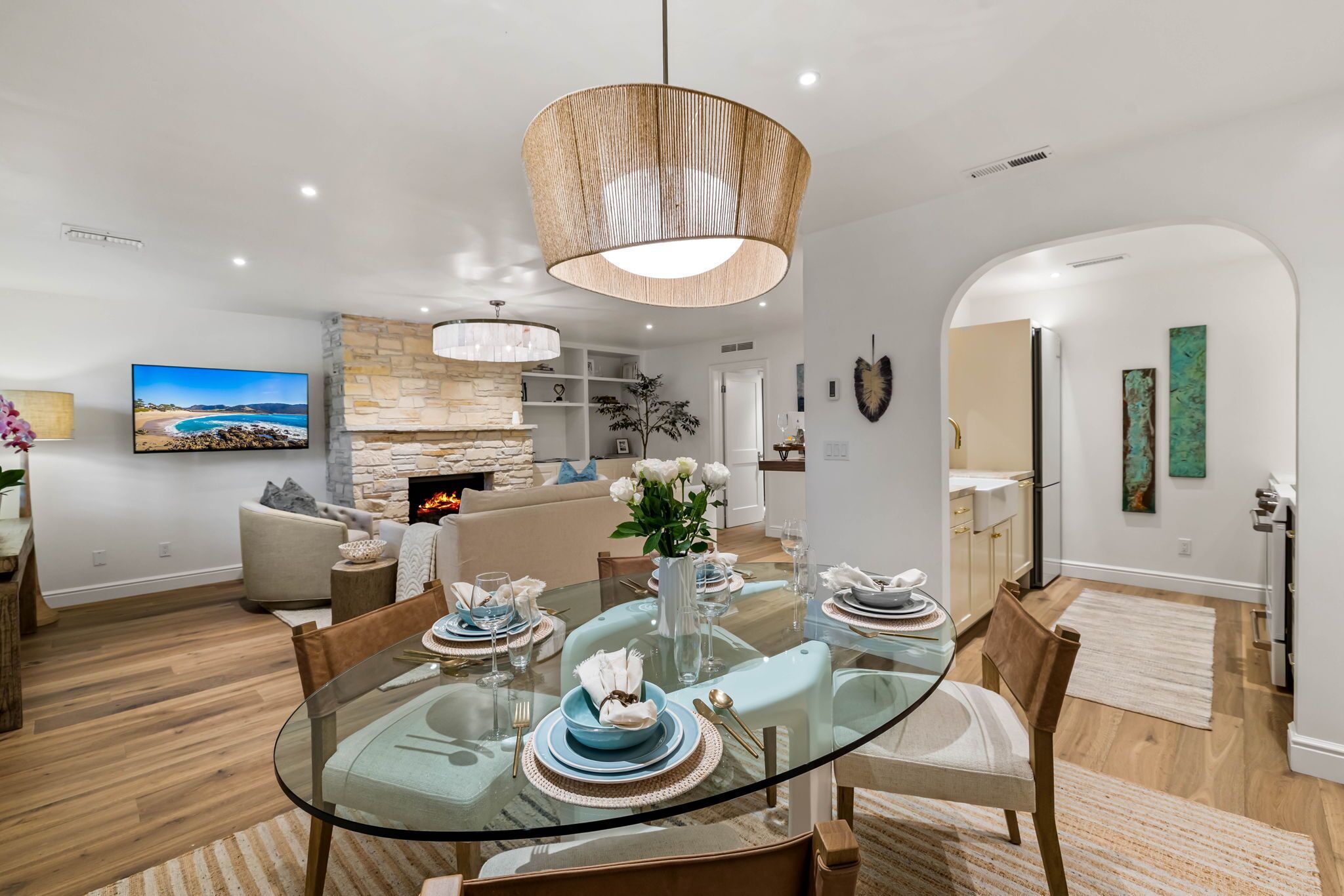 A modern dining area with a glass table, wooden chairs, and a central hanging light fixture. A stone fireplace and a television are visible in the background.