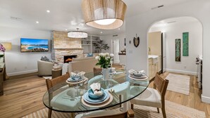 A modern dining area with a glass table, wooden chairs, and a central hanging light fixture. A stone fireplace and a television are visible in the background. - 3972 DolceVita ~Beautiful Remodel, Walk Everywhere (Carmel)
