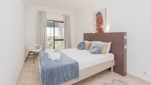 A bedroom with a bed, a chair, a painting, and a view of the outside. - Central Apartment - Rooftop pool (Albufeira)