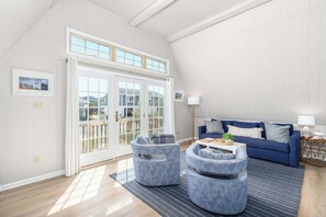 Un salón amplio con techo alto, un sofá azul, dos sillones azules, una mesa de centro y un ventanal con vistas a casas.