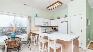 A modern kitchen with white cabinets, a large island with bar stools, a window with a view, and a ceiling light fixture. - Waterfront Condo with Pool (Ocean City)