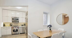 Une cuisine moderne avec des meubles blancs, des appareils en acier inoxydable et un coin repas comprenant une table en bois et des chaises blanches.