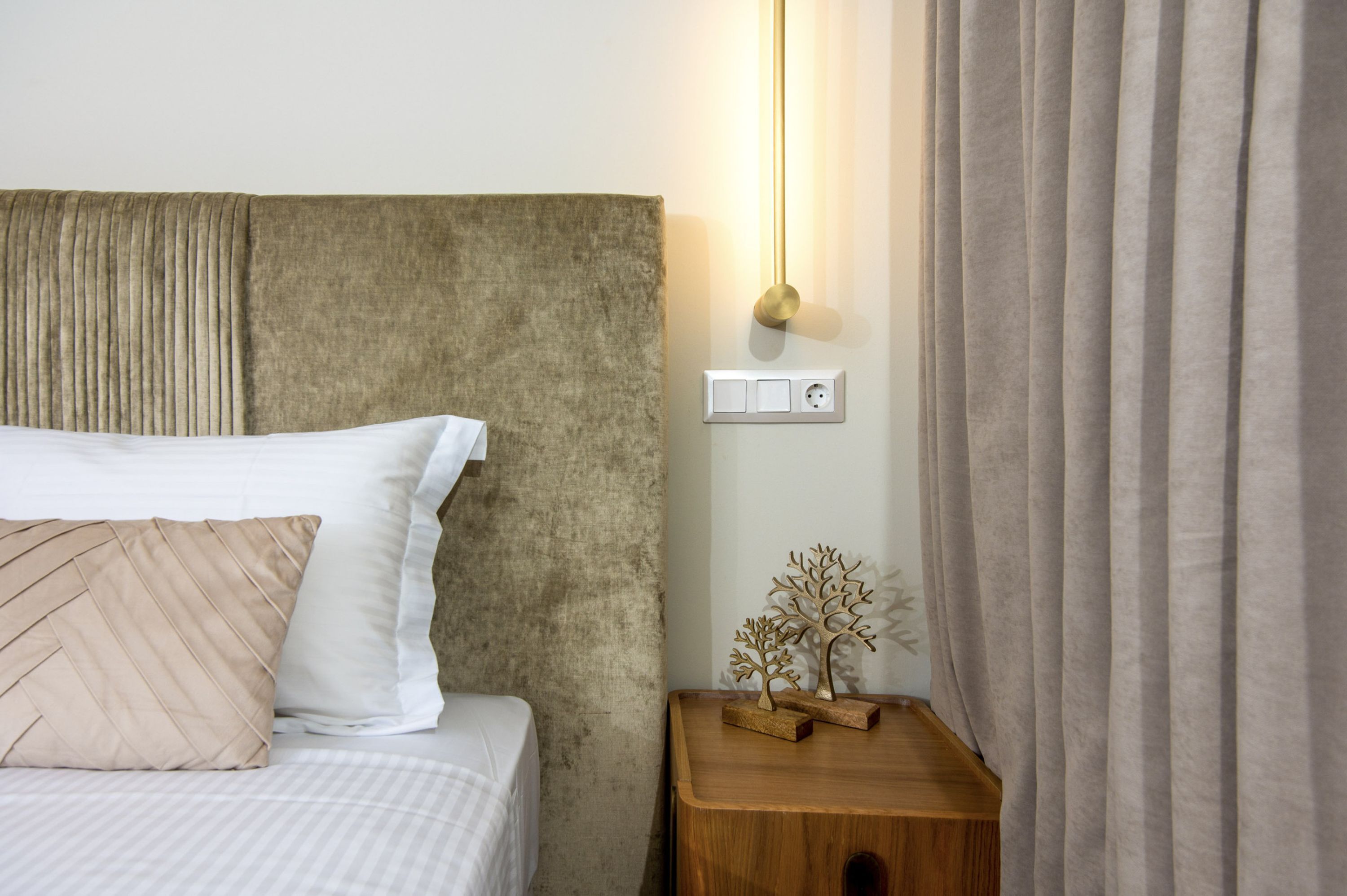 A neatly made bed with white and beige pillows, a wooden nightstand with a small tree decoration, and a wall-mounted light fixture.