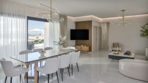 A modern living room with a glass dining table, white chairs, a flat-screen TV, and a large window with curtains.