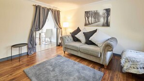 Una sala de estar con un sofá gris, una mesita redonda pequeña, una alfombra gris y un cuadro de árboles en la pared.