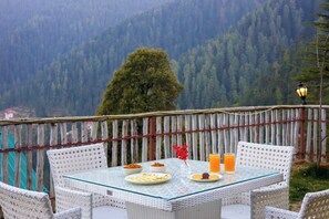 A table set with plates, bowls, and a glass of orange juice, surrounded by wicker chairs, with a view of a forested mountain range in the background. - Alaya Stays Luuma Luxury Villa - Hillside 5-BHK Villa Amidst Pines With Garden, Balcony & Lounge (Shimla)
