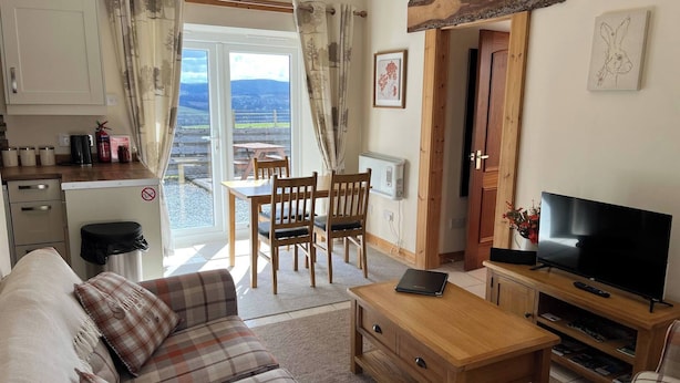 A cozy living room with a sofa, a coffee table, and a TV. There's a dining area with chairs and a table, and a kitchen area with a fridge and a kettle. - Fabulous Open Views Over Dufftown (Keith)