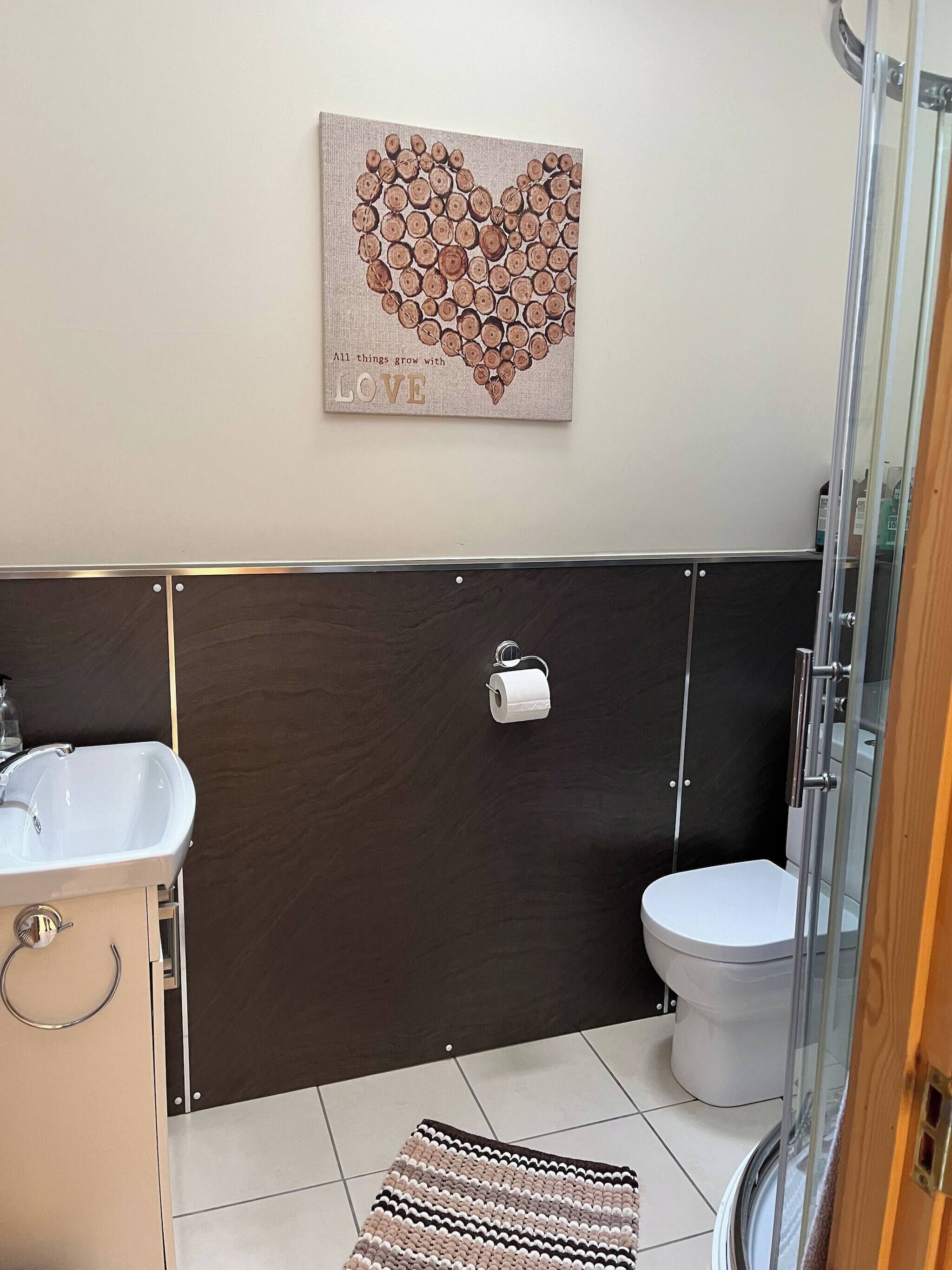 A bathroom with a white sink, toilet, and shower. There's a heart-shaped artwork on the wall and a striped rug on the floor.