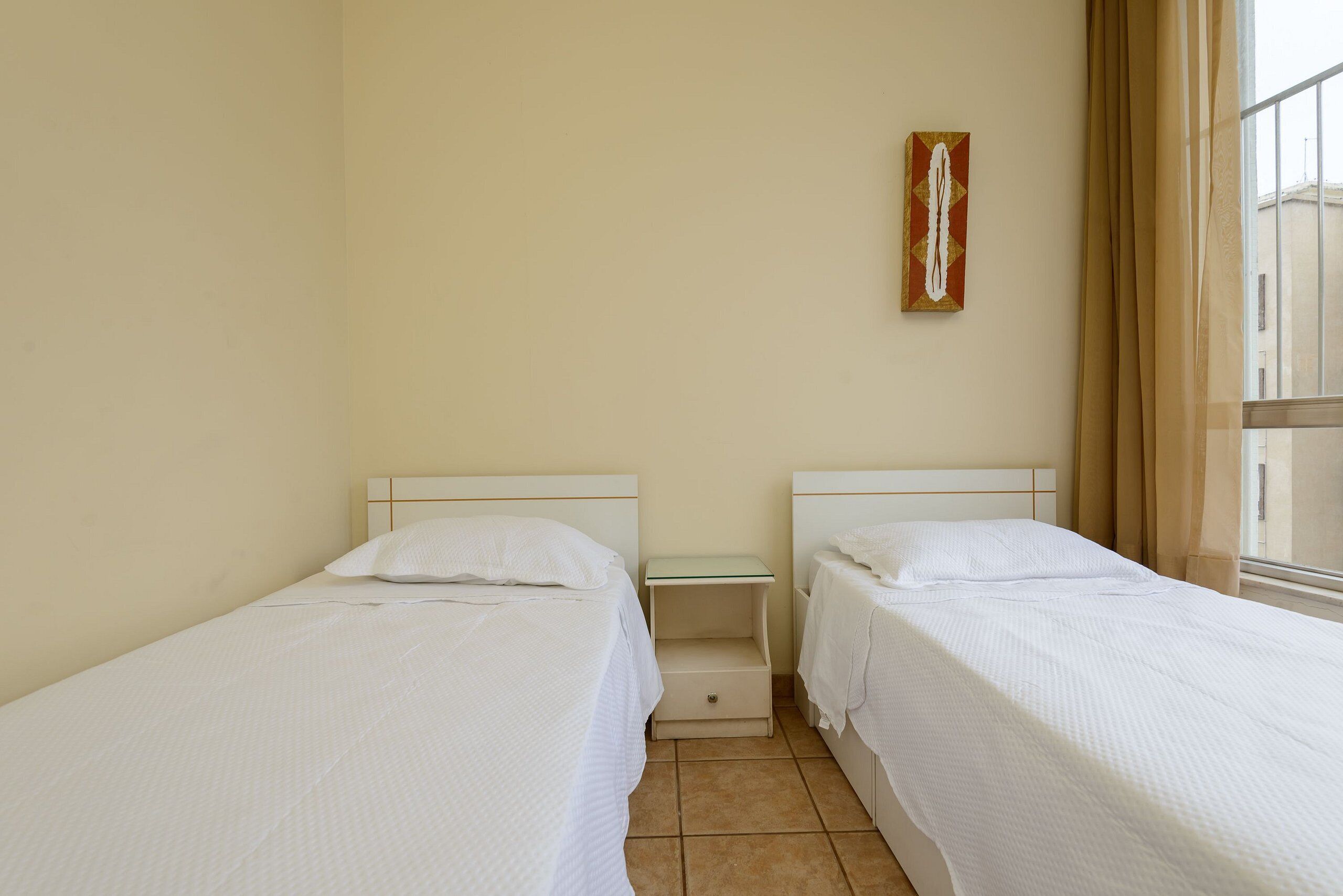 Une chambre avec deux lits simples, une petite table de chevet, une œuvre d’art accrochée au mur et une fenêtre avec des rideaux.
