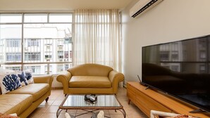 A modern living room with a sofa, armchair, and TV, featuring a large window with curtains and a city view. - Rj04 Ap em Copacabana até 6 pessoas (Rio de Janeiro)