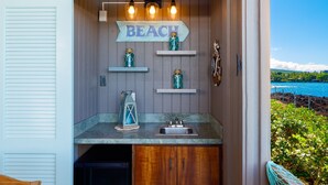 A small kitchen with a sink, shelves, and a 'BEACH' sign. - Big Island Kanaloa At Kona 2802 2 Bedroom Condo (Kailua-Kona)