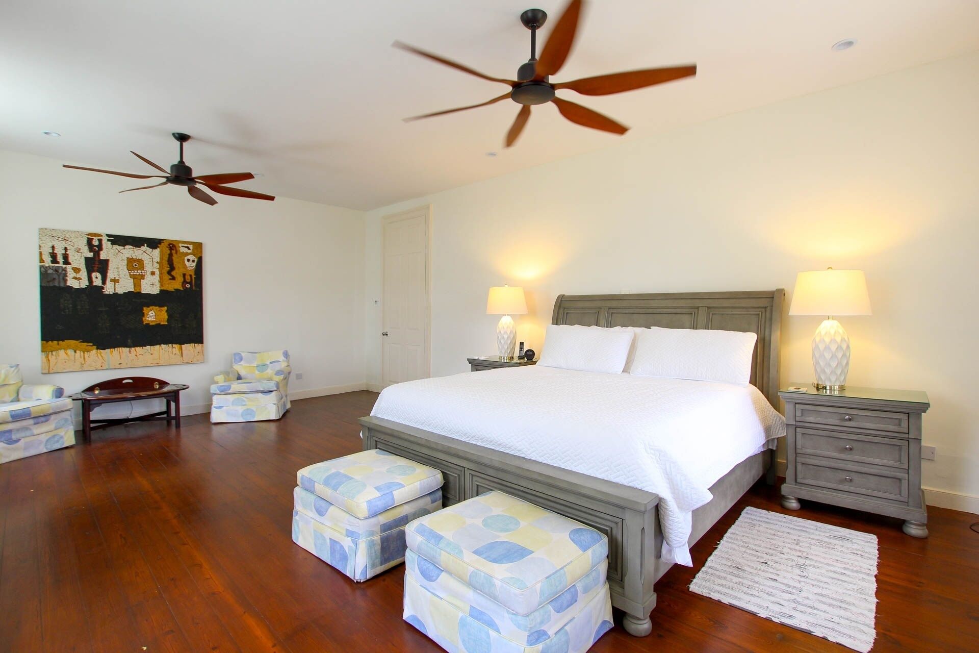 A bedroom with a large bed, two nightstands, a ceiling fan, a painting, and a wooden floor.