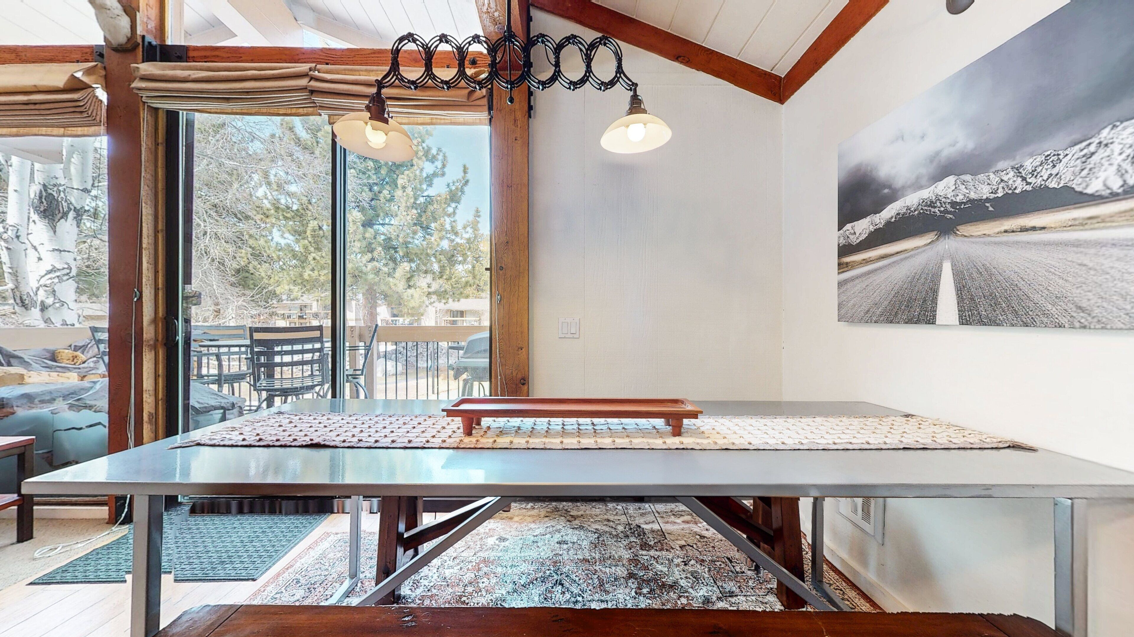 A dining area with a large table, two hanging pendant lights, and a mountain landscape painting on the wall.