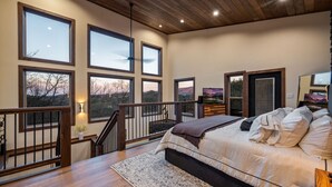A spacious bedroom with a large bed, multiple windows offering views of the outdoors, a staircase, and a wooden ceiling. - Reflections 1 Bedroom Cabin (Sevierville)