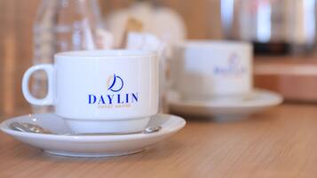 A white cup with "DAYLIN GUEST HOUSE" logo on a saucer, a clear water bottle, and a silver coffee pot on a wooden table.