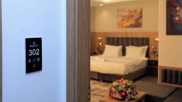 A hotel room with a bed, a nightstand, a bedside table with a flower arrangement, and a wall-mounted control panel displaying room number 302.