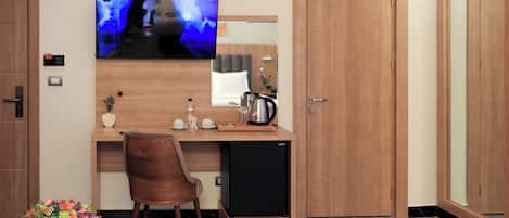 A compact hotel room with a wooden desk, a television mounted on the wall, a small refrigerator, and a wooden wardrobe.