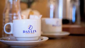 A DAYLIN branded cup and saucer on a wooden table with a blurred background featuring a coffee pot and a water bottle.