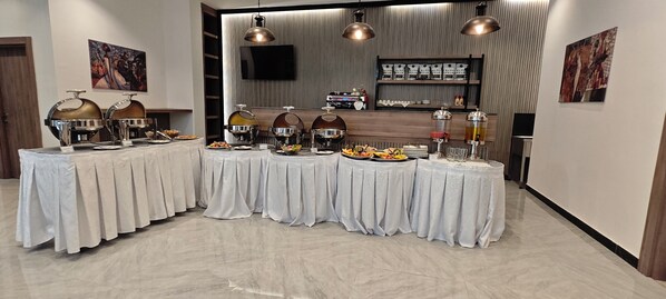 A buffet setup with multiple round tables covered in white tablecloths, each with food and drink dispensers.