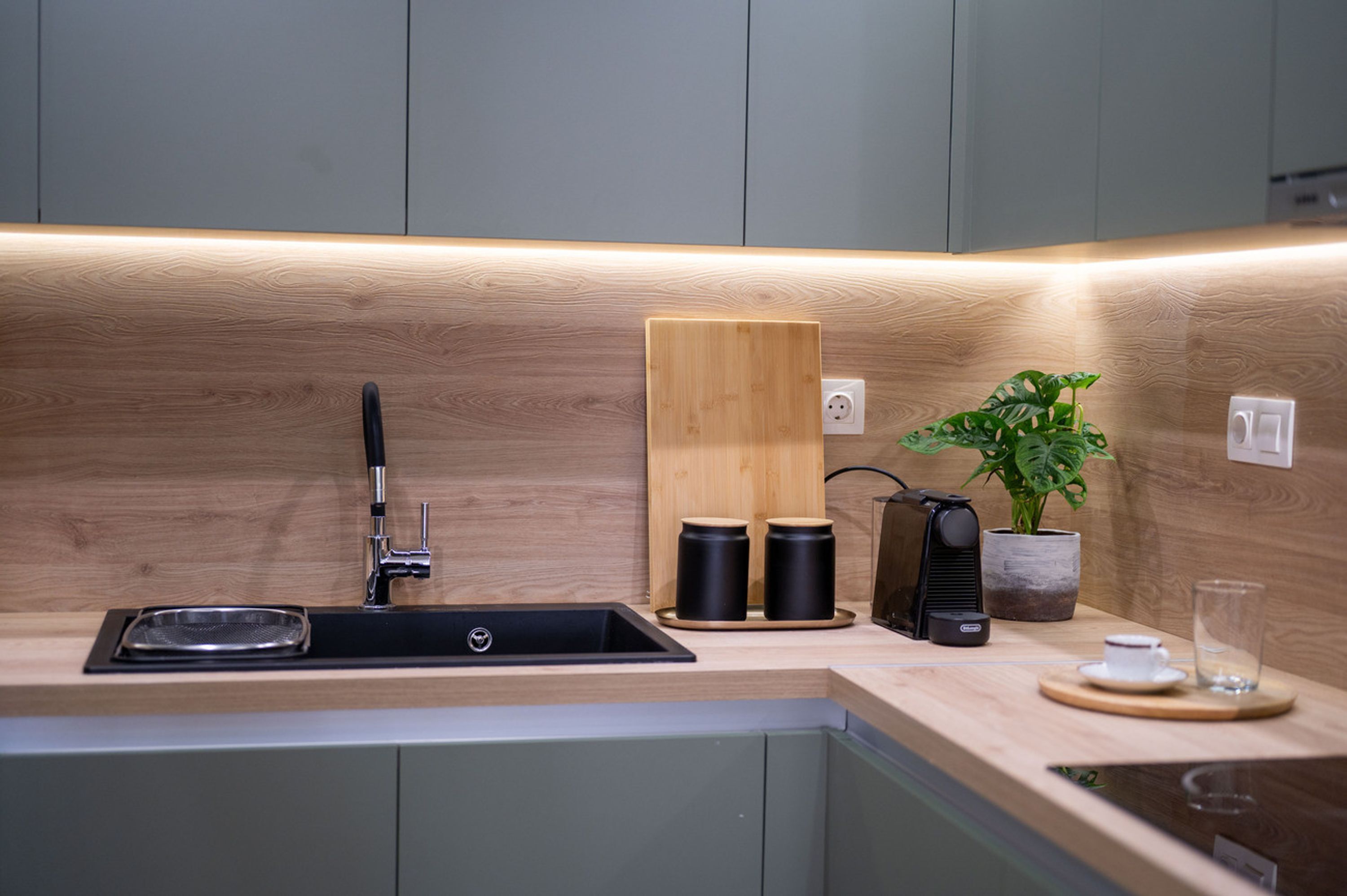 A modern kitchen with a wooden countertop, a sink, a cutting board, two black containers, a coffee maker, a potted plant, and a glass.