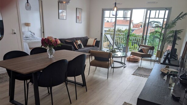 A modern living room with a wooden dining table, black chairs, a sofa, and a balcony with a railing. - Appartement au coeur dándernos les Bains (Andernos-les-Bains)