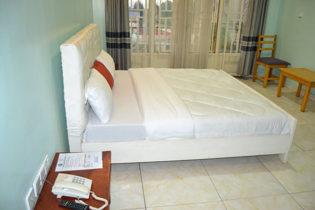 A hotel room with a bed, a wooden chair, a small table with a telephone, and a window with curtains.