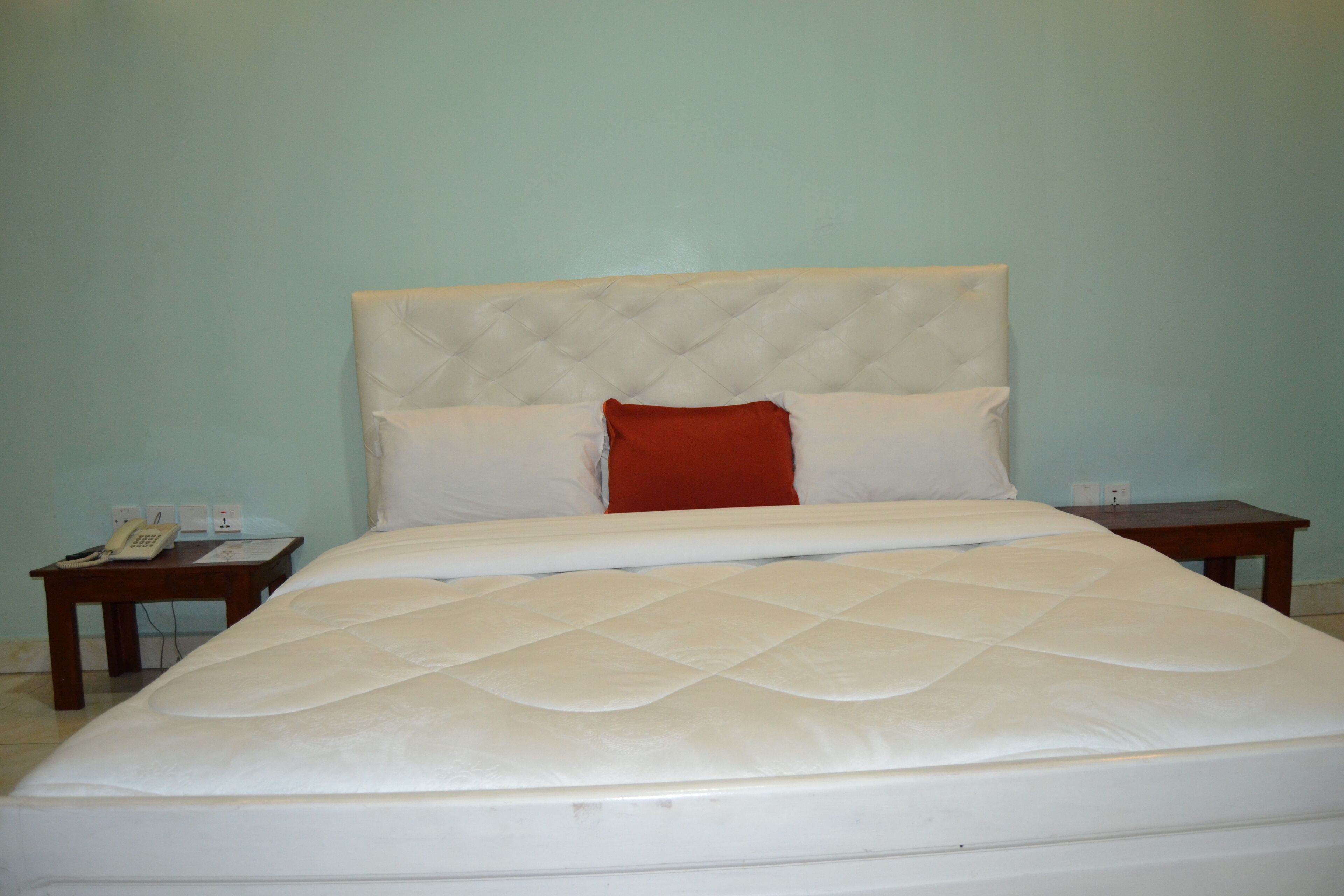 A neatly made bed with a white headboard and a red accent pillow, a bedside table with a phone and a lamp, and a wall socket.