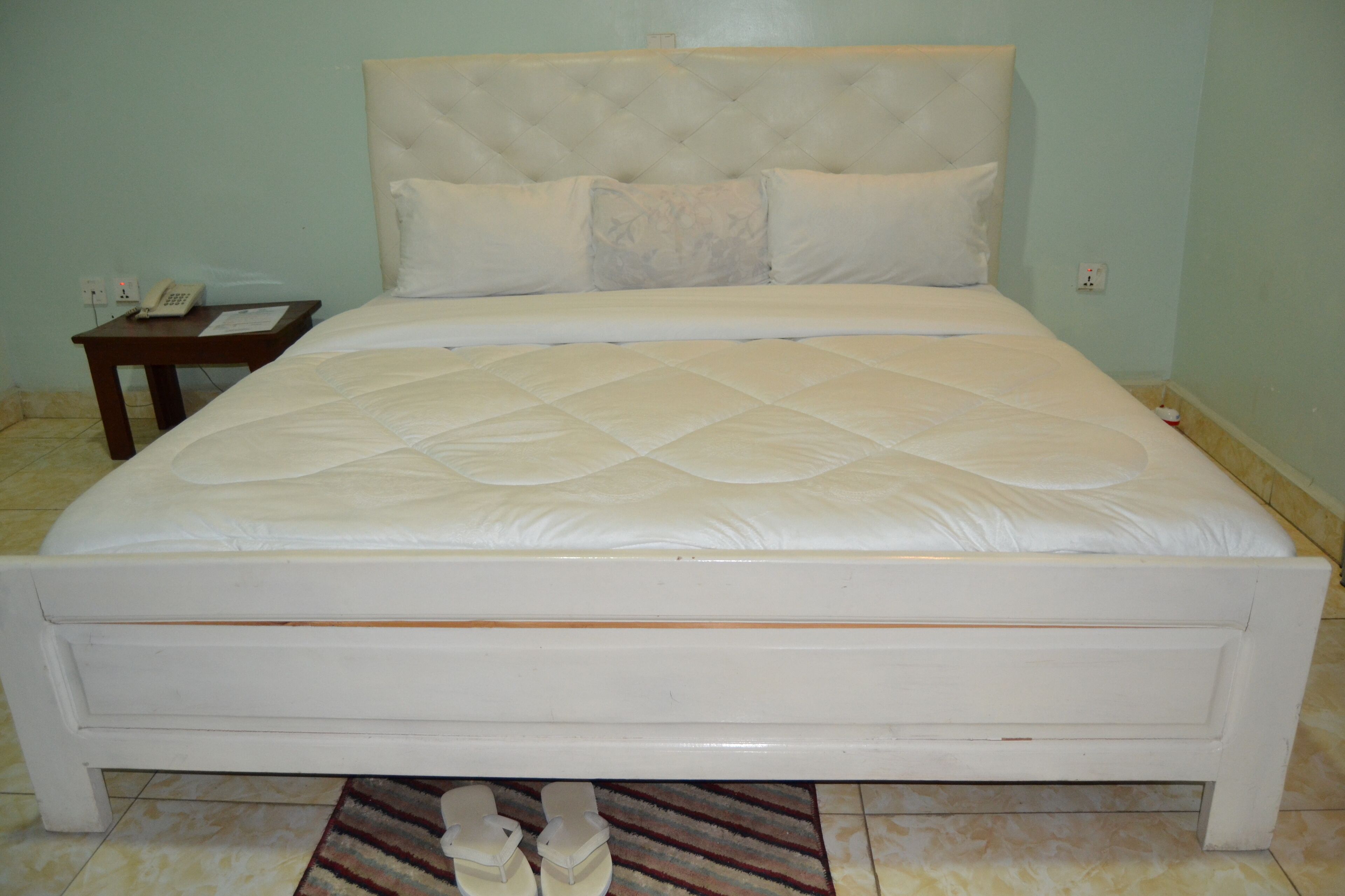 A white bed with a quilted mattress and a headboard, a small wooden nightstand with a telephone, and a striped rug on the floor.