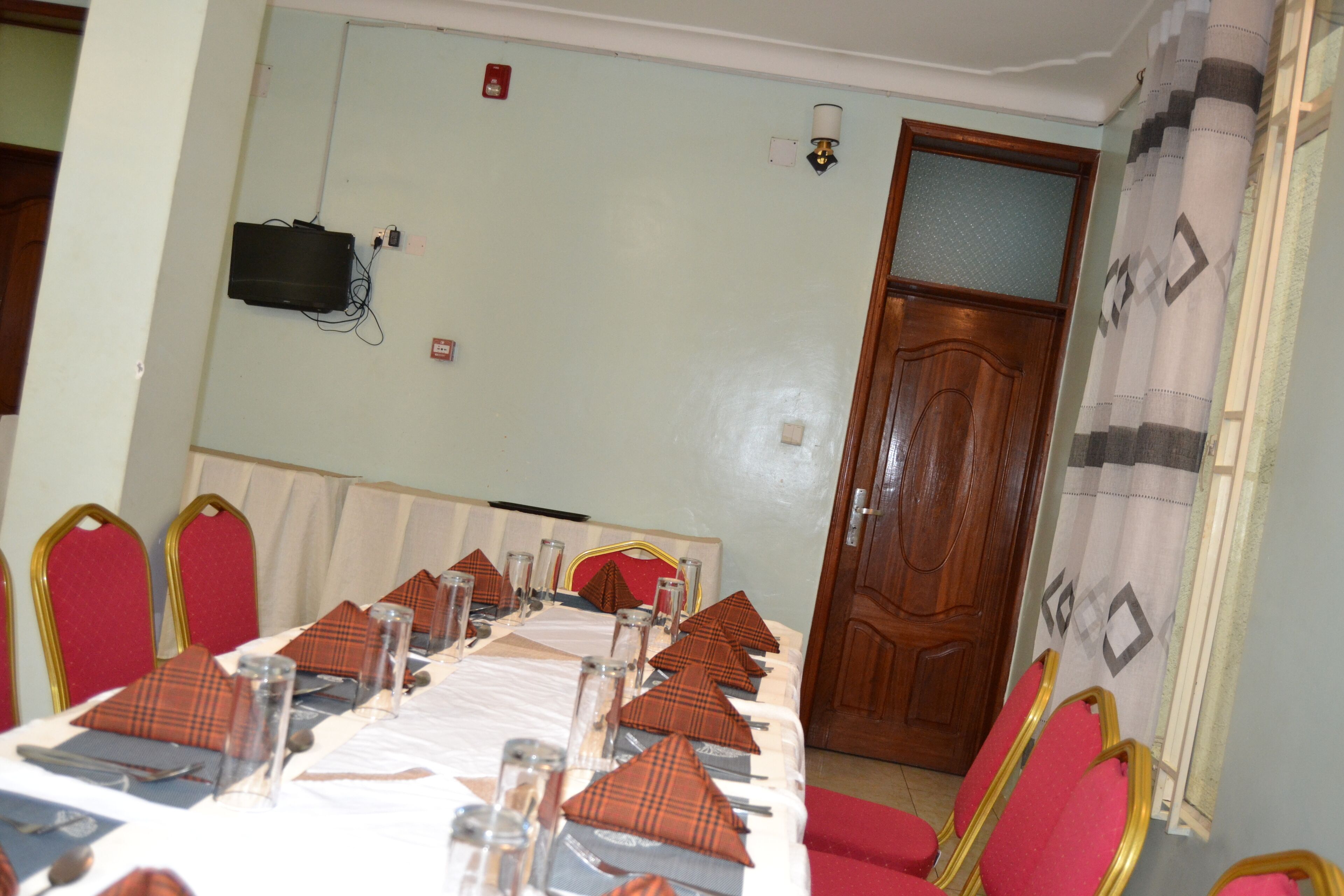 Un salón de conferencias con una larga mesa dispuesta para una reunión, sillas rojas, una puerta de madera y un proyector montado en la pared.