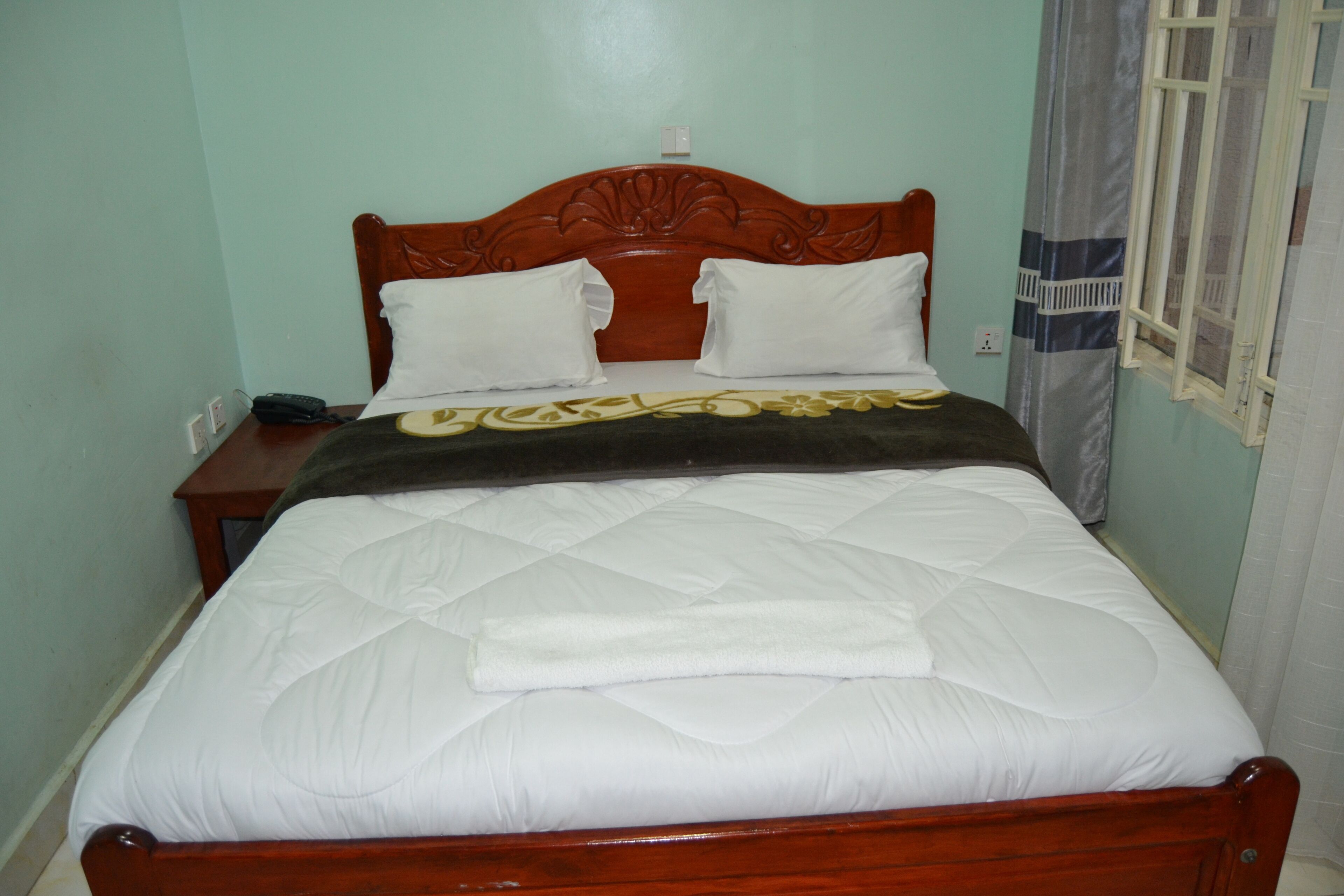 A neatly made bed with a wooden headboard, a bedside table with a phone, and a window with a curtain.
