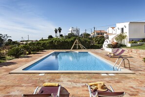 A rectangular swimming pool with a clear blue water surface, surrounded by a brick patio. There are lounge chairs with towels and hats placed on the patio. - Quinta dos Medronheiros (Castro Marim)