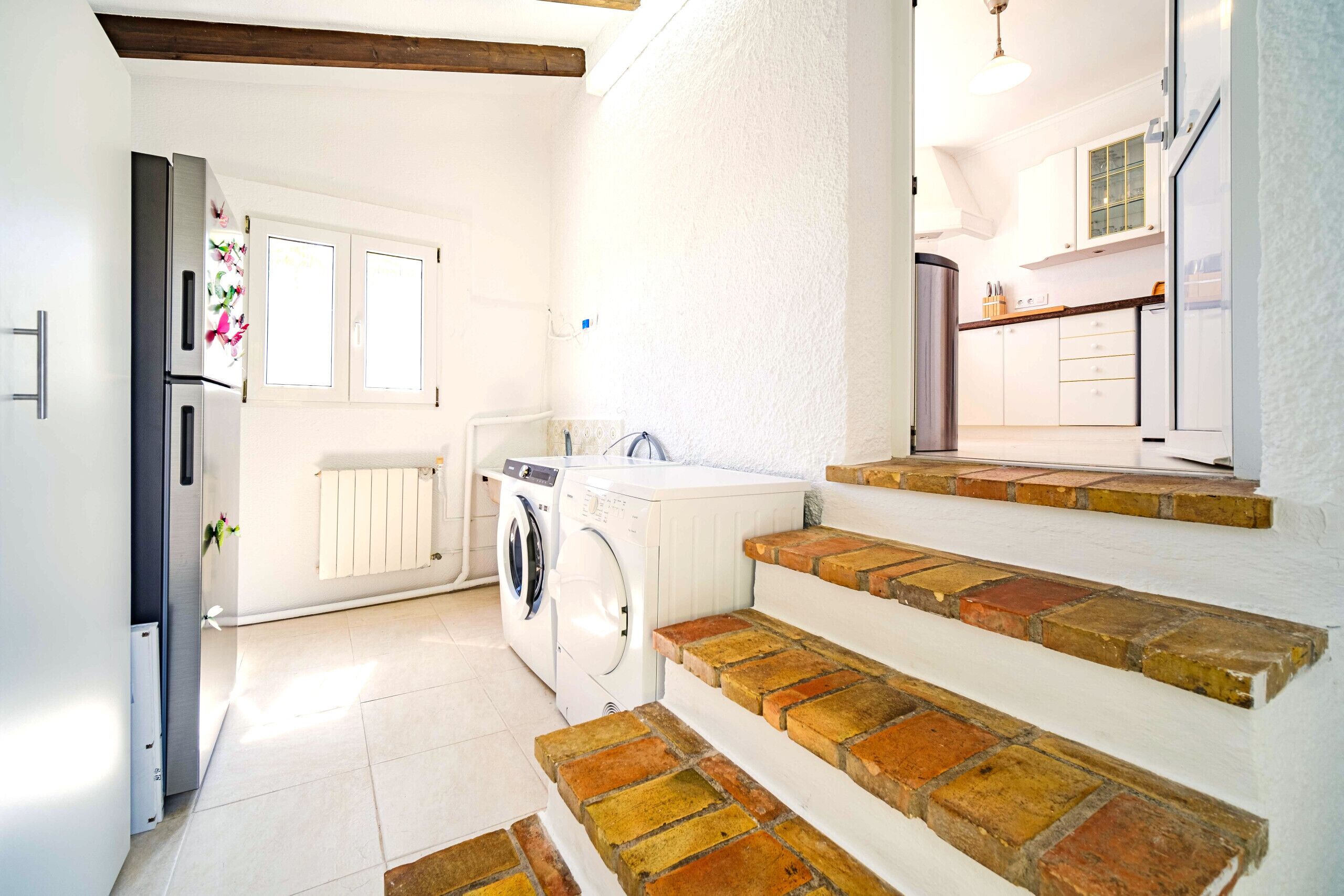 A laundry area with a washing machine, dryer, and a radiator. There are steps leading to a kitchen area with white cabinets and a countertop.