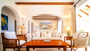 Amplia sala de estar con una mesa de centro de madera, sofás blancos y sillas de mimbre. Hay dos cuadros en la pared, una lámpara a cada lado, y vista al mar a través de la ventana.