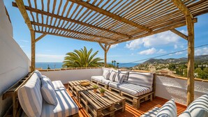 Una terraza con pérgola de madera, con un sofá y una mesa de centro, con vista a un paisaje costero.