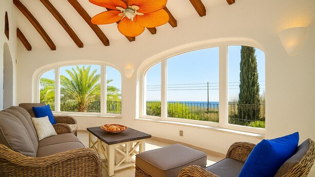 Una habitación iluminada por el sol, con muebles de mimbre, una mesa con un cuenco y ventanas amplias que ofrecen vistas a la vegetación y un paisaje lejano.