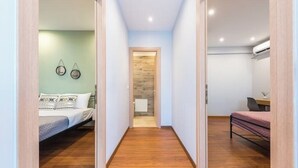 A narrow hallway with wooden flooring leading to a bedroom with a bed and a desk.